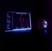 Installation in a dark room showing two screens set up next to each other displaying a webpage interface with a menu column and a human body in between white text on the left and the back of a body from the head down to butt on the right.