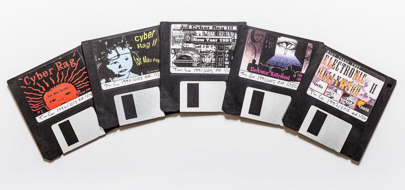 Five floppy disks fanned out on a white surface. Each have color-printed graphic collaged images, evoking anarchy and rebellion with pictures of baby faces, a motherboard, a movie theater with an eye melting on the screen, and a factory burning in flames.